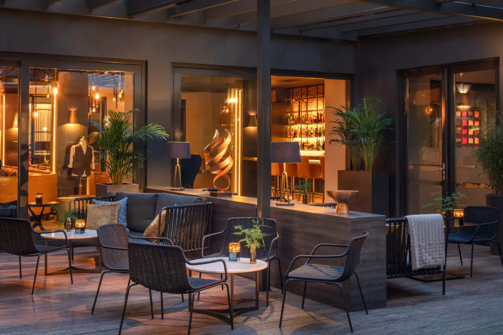 De verwarmde patio van Bar The Tailor bij avond, met rieten stoelen, kaarslicht en een etalagepop in maatpak achter het raam