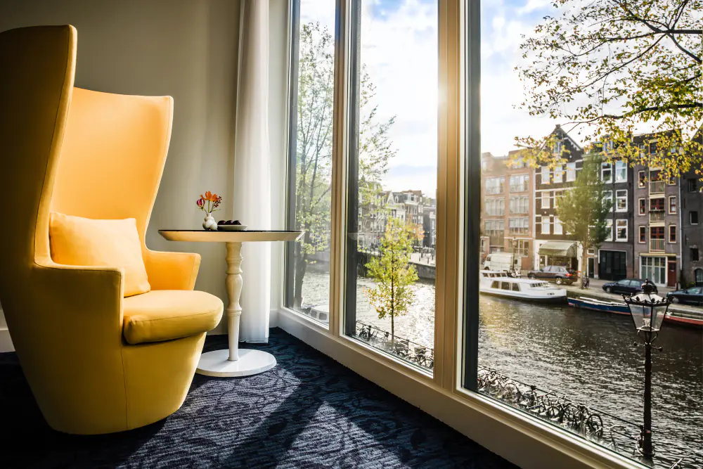 Een hotelkamer met een gele fauteuil bij hoge ramen en uitzicht op de gracht