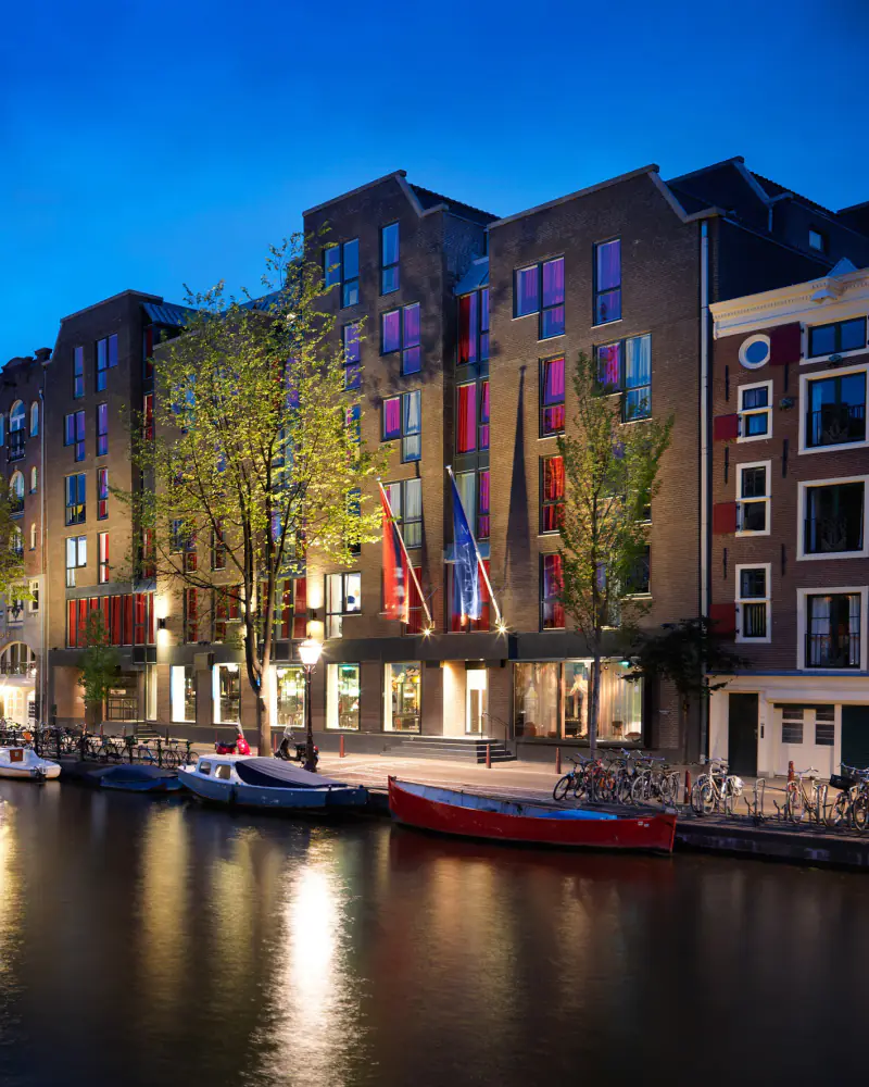 Grachtenpanden bij avond, met roze en paarse ramen, vlaggen en bootjes op het water
