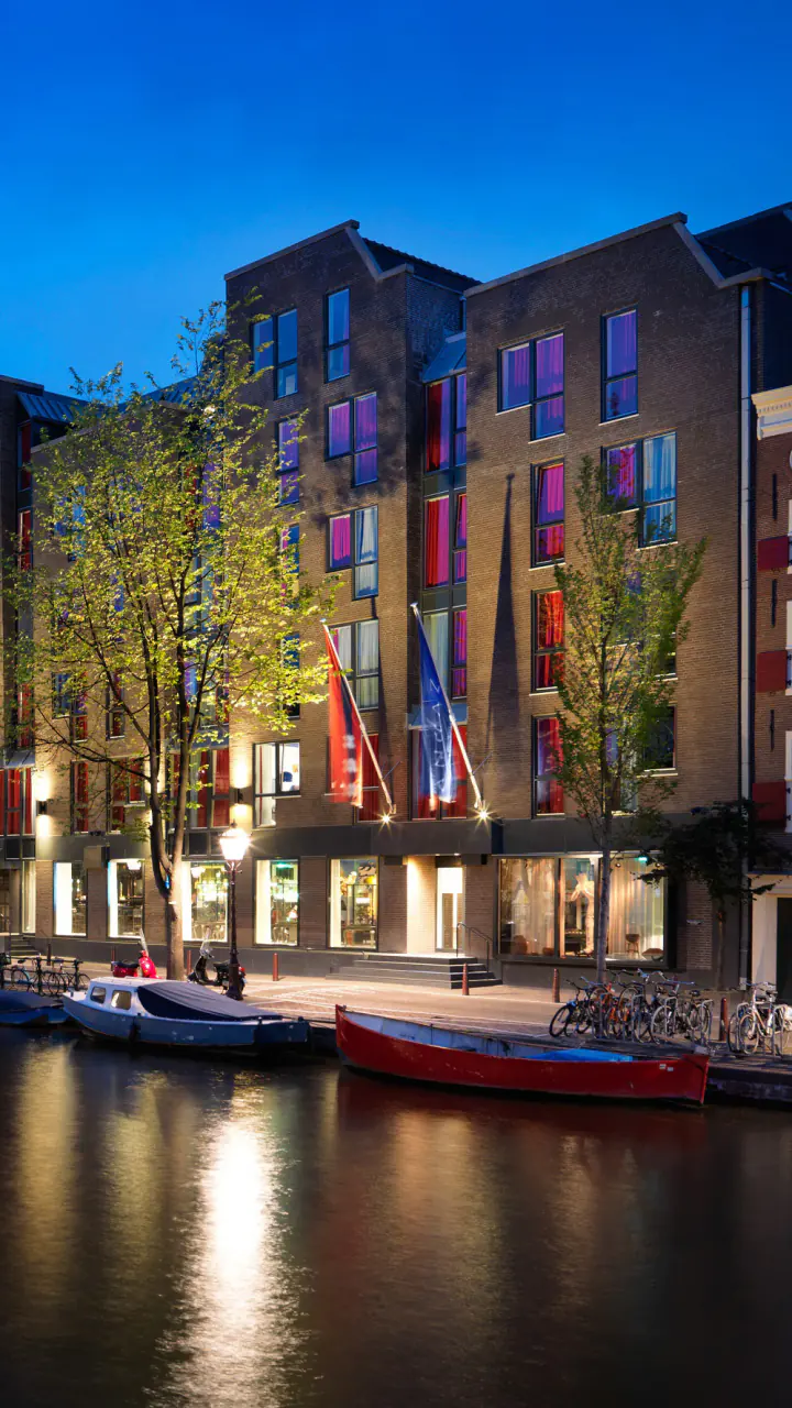 De grachtenpanden van Andaz Amsterdam Prinsengracht bij avond, met roze en paarse ramen, vlaggen en bootjes op het water