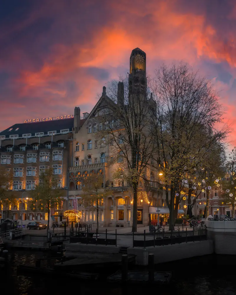 De klokkentoren aan de Leidsekade bij zonsondergang, met lichtletters op het dak en lichtjes in de bomen