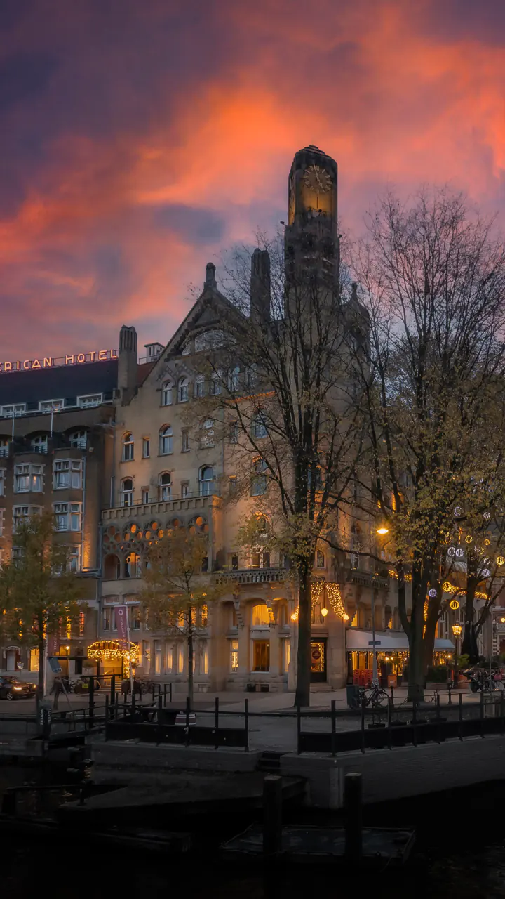 Het American Hotel aan de Leidsekade bij zonsondergang, met de klokkentoren, lichtletters op het dak en lichtjes in de bomen
