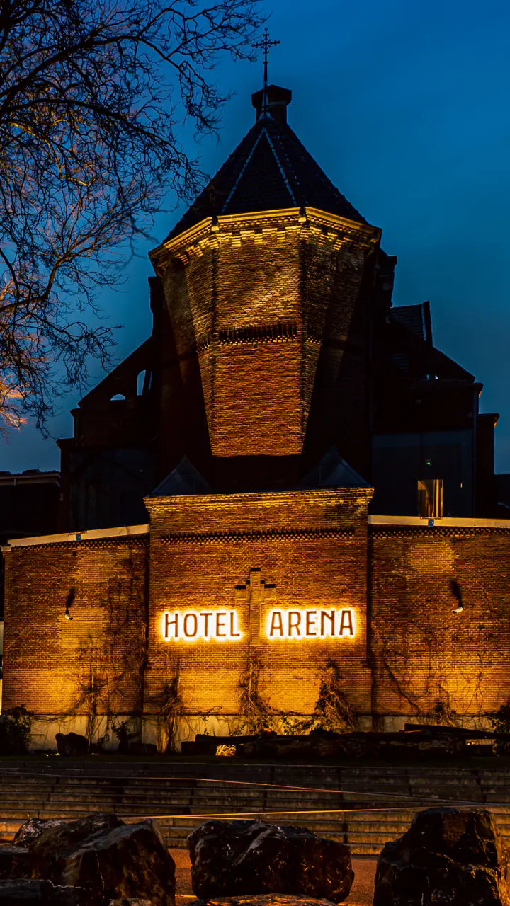 De verlichte bakstenen kapel van Hotel Arena bij avond, met de lichtletters Hotel Arena en kale bomen