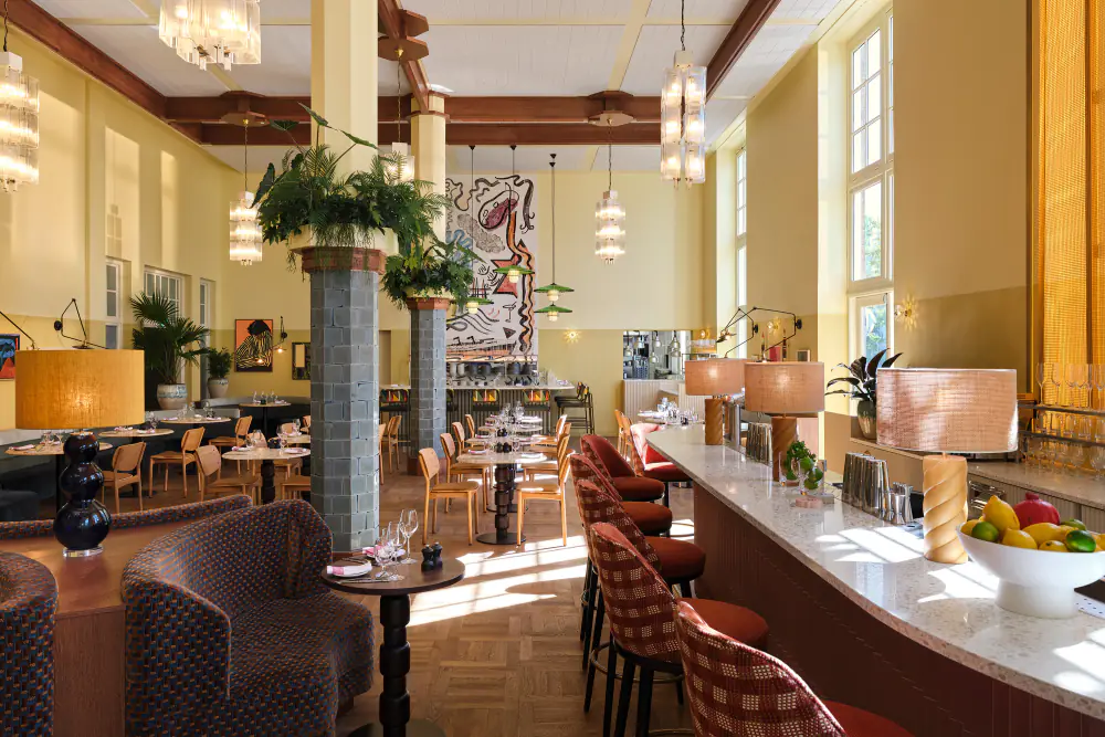 De zaal van Breman Brasserie met hoge ramen, zuilen, planten en een lange bar