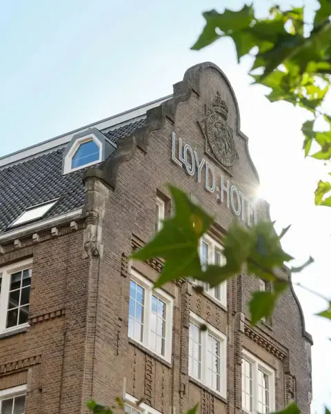 De bakstenen trapgevel met de letters Lloyd Hotel en een wapen, tussen groene bladeren in de zon