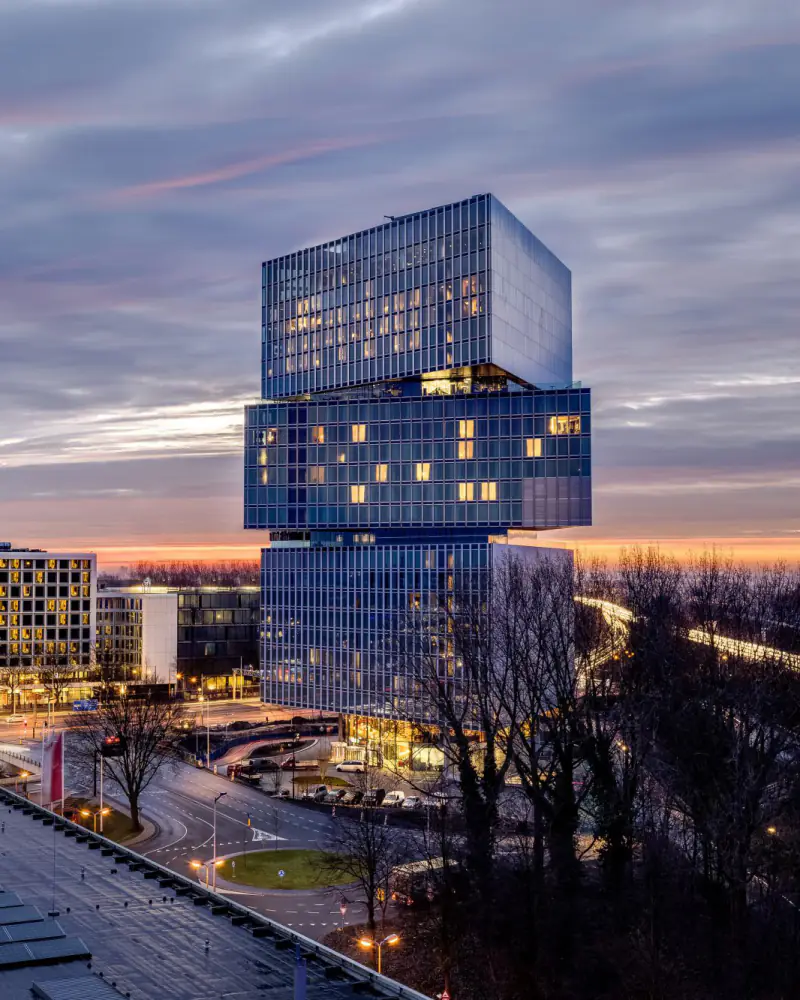 De gestapelde glazen toren in de schemering, met verlichte ramen en de ringweg ernaast
