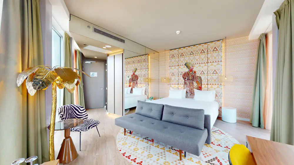 Een hotelkamer van nhow met een bed, een grijze bank, een gouden lamp en een schilderij boven het bed