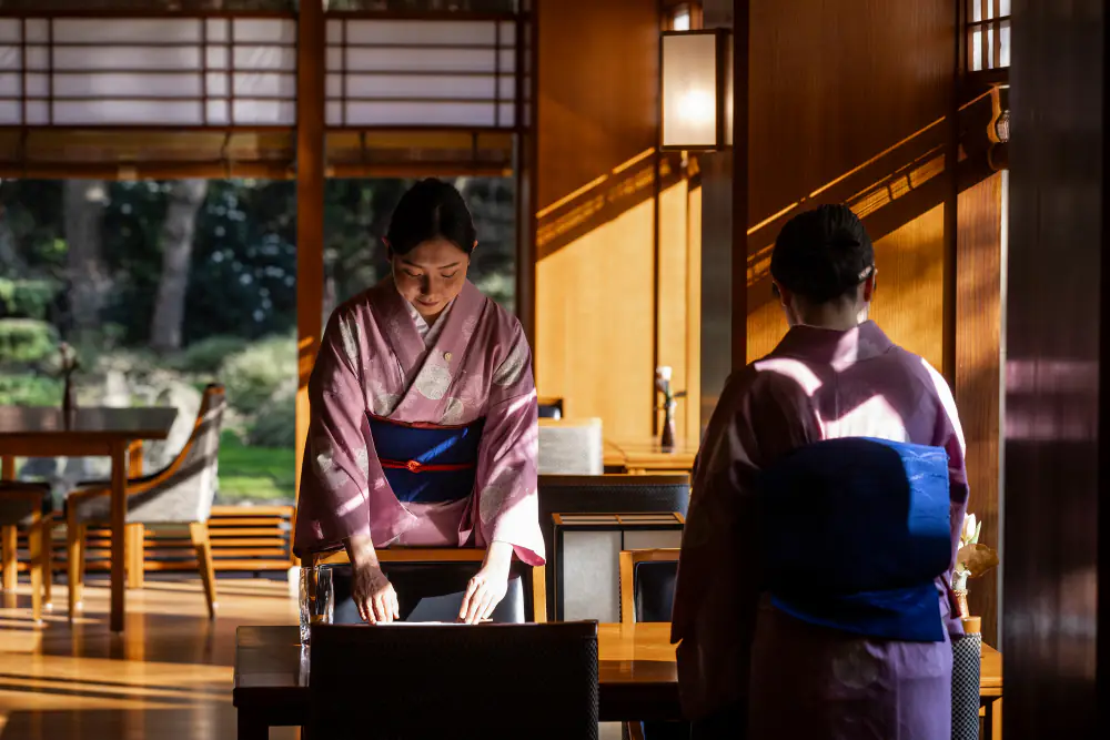 Twee medewerkers in kimono dekken een tafel in Yamazato, met de tuin achter de ramen