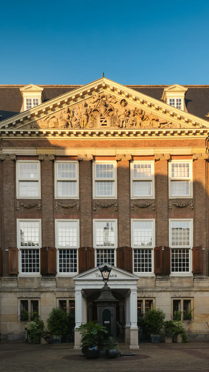 De gevel van The Grand aan de binnenplaats in de avondzon, met een gebeeldhouwd fronton, luiken en windvanen