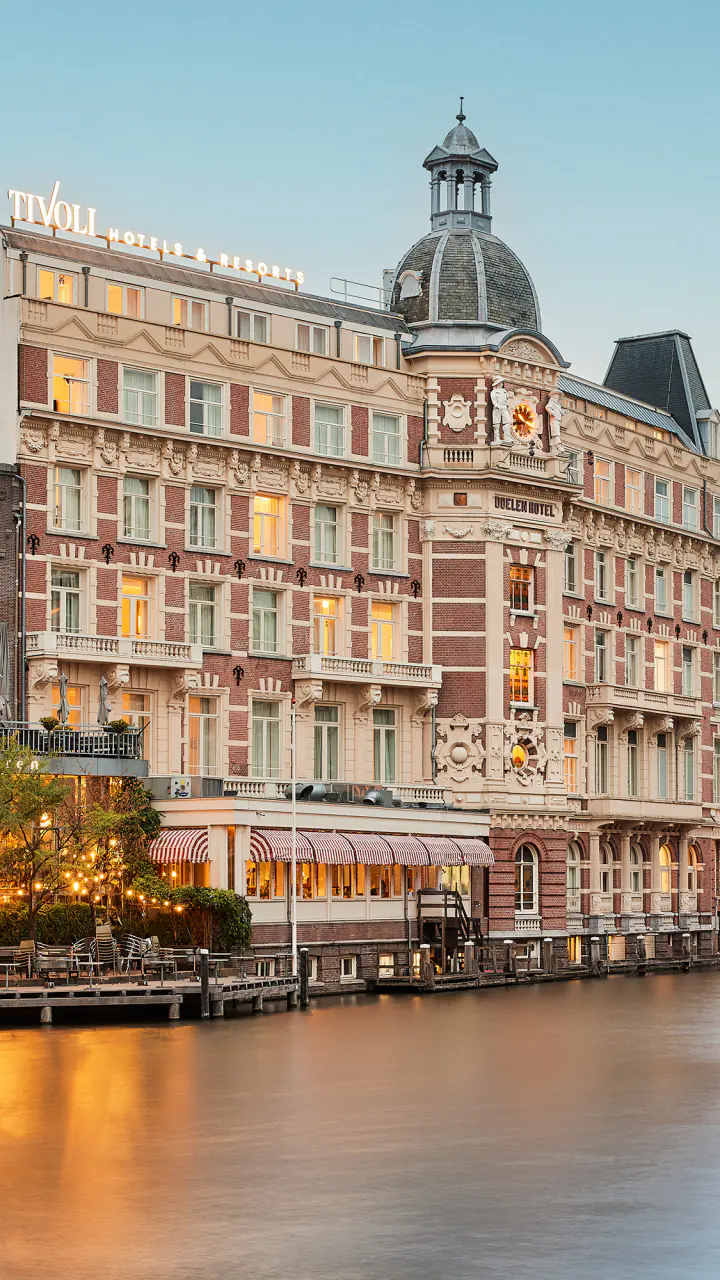 Tivoli Doelen aan het water bij schemering, met een koepeltorentje, verlichte ramen en een verlichte brug op de voorgrond