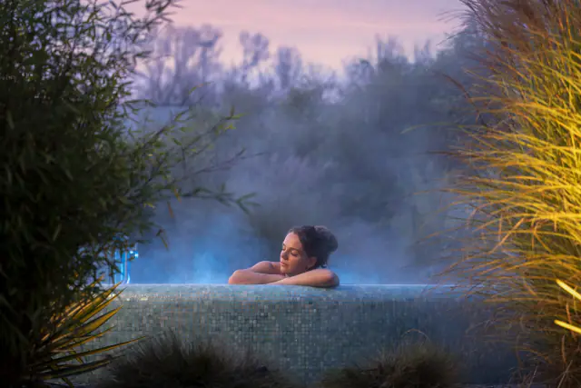 Een vrouw in een dampend buitenbad van Zuiver in de schemering, tussen het groen