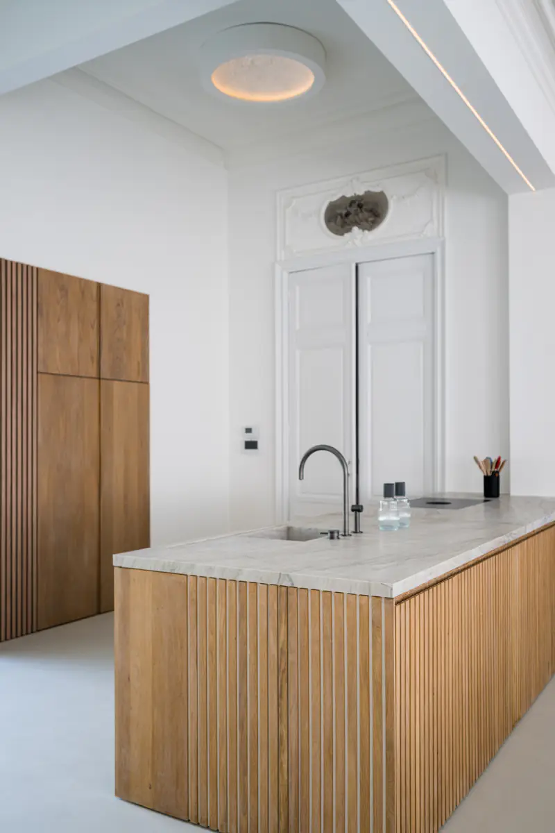 Kitchen island with oak slats and a marble top, with a dark tap, in a white room with ornaments above the door