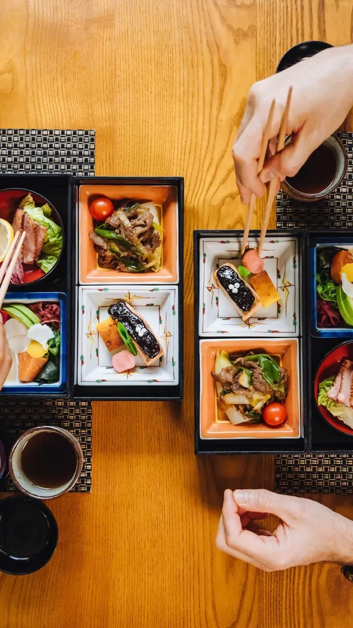 Twee bentoboxen van KIRI van bovenaf, op een houten tafel, met handen die met stokjes eten